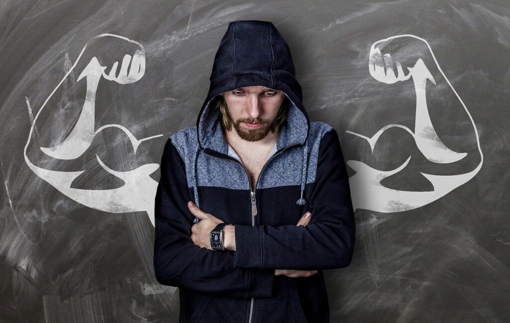 A man with folded arms standing in front of a blackboard with a drawing of muscular arms for strength