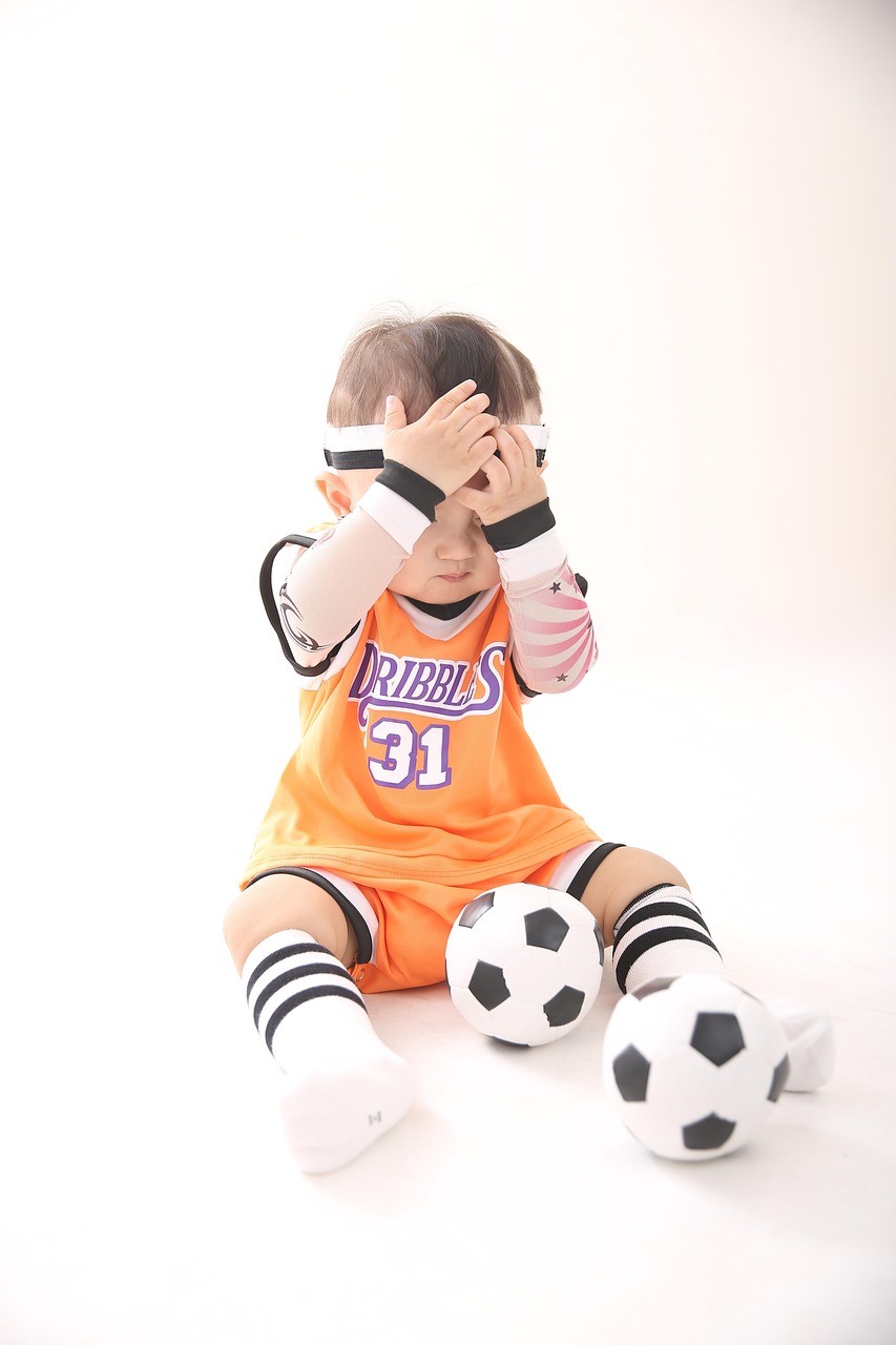 white baby boy sitting with two footballs between his legs; covered eyes