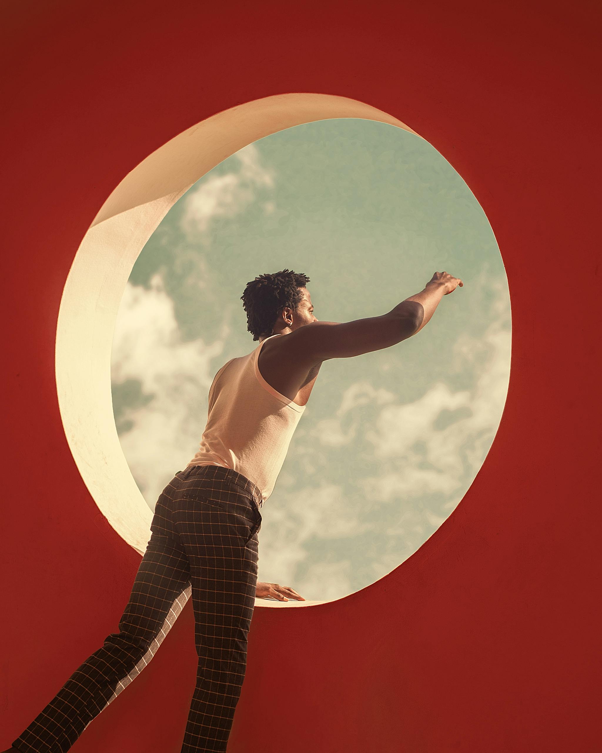 A man in a tank top reaches through a round window towards the sky, against a red backdrop.