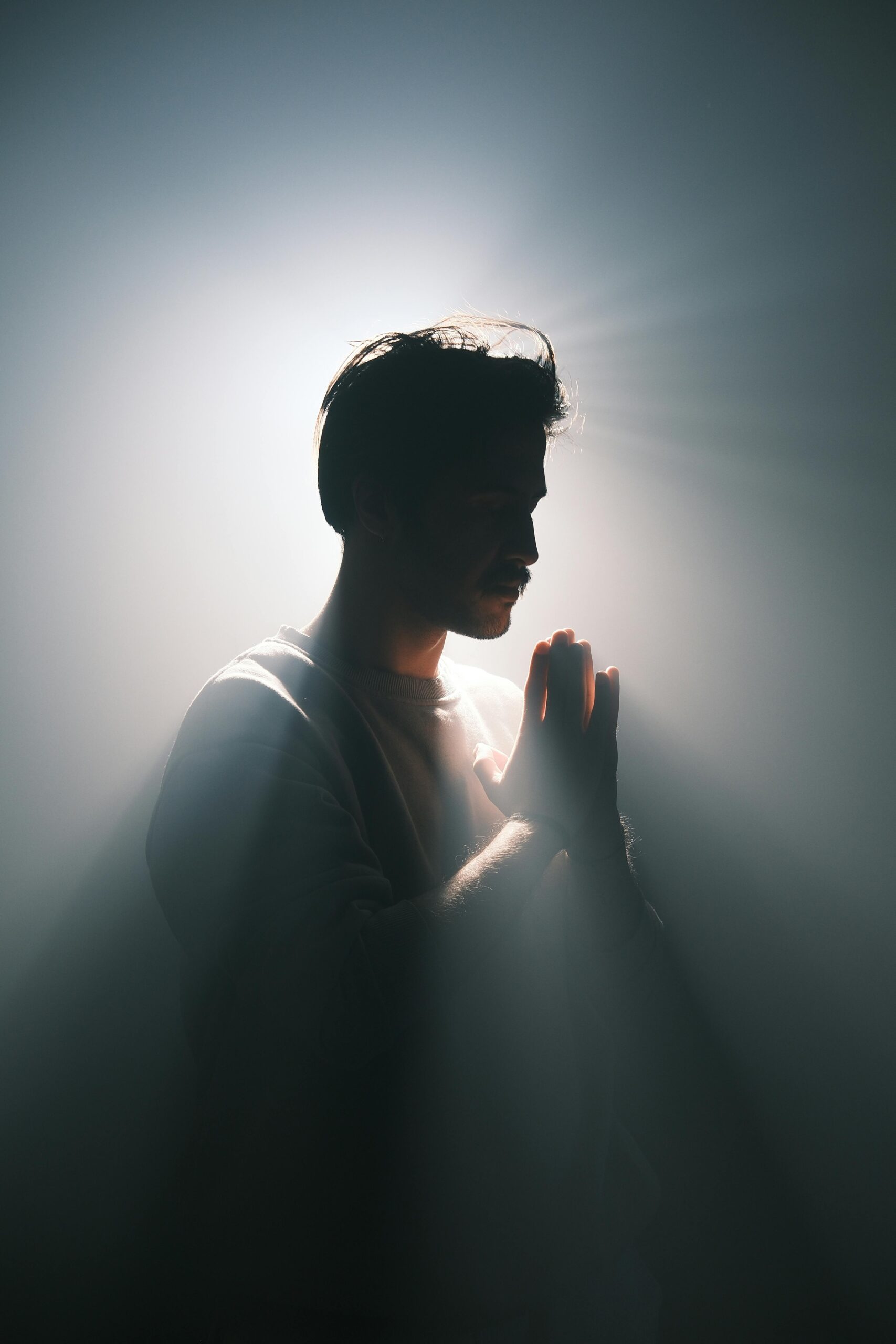 A silhouette of a man in prayer with dramatic backlighting creating a halo effect and light rays.