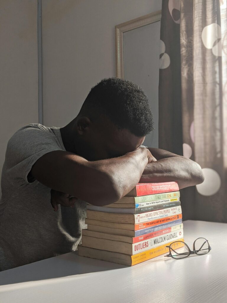 A young man rests on a stack of books at a desk, sunlight filtering through a window.