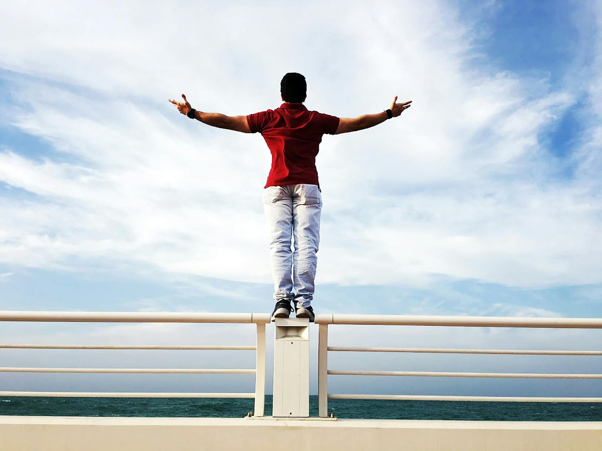 A person standing on a railing by the sea with arms outstretched under a clear sky, symbolizing freedom.
