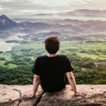A man sitting on a cliff, appreciating a breathtaking mountain landscape in daylight.