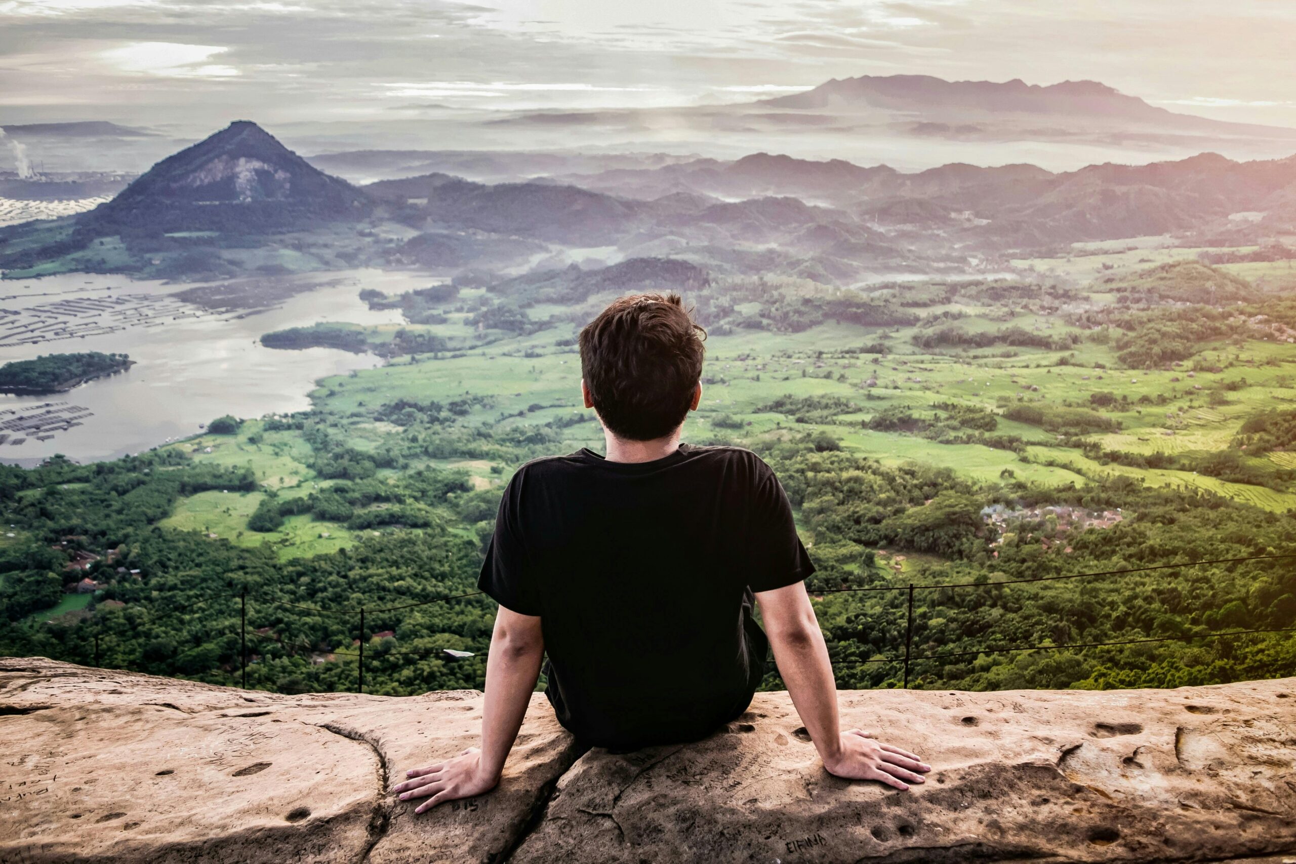 A man sitting on a cliff, appreciating a breathtaking mountain landscape in daylight.
